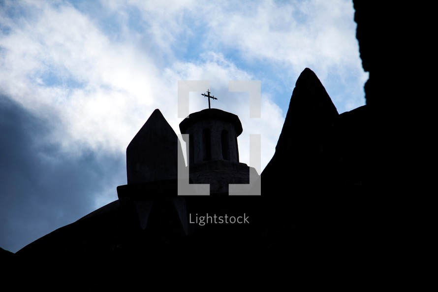 Silhouette of church steeple with cross on top with blue sky and clouds in the background.