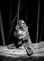 woman sitting on fallen tree wearing a fur coat and holding her legs