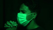 man standing in green light wearing a surgical mask and praying 