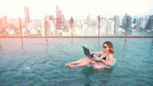 A young entrepreneur woman is working on her computer in by the resort pool in summer time. Beautiful young female model working freelance at swimming pool on laptop.	