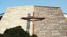 A wooden cross on a brick wall with a green bush in front of a church with bright shining light.