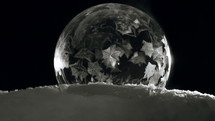 Ice crystals forming in slow motion on freezing bubble sitting on snow in front of black background.