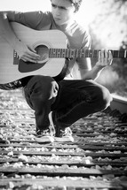 teen boy playing a guitar standing on train tracks 