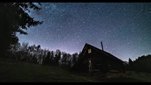 Time lapse of Starry night sky with milky way galaxy rotates fast over old wooden hut in wild forest.