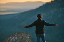 a man with outstretched arms standing on a mountaintop looking out at the view 