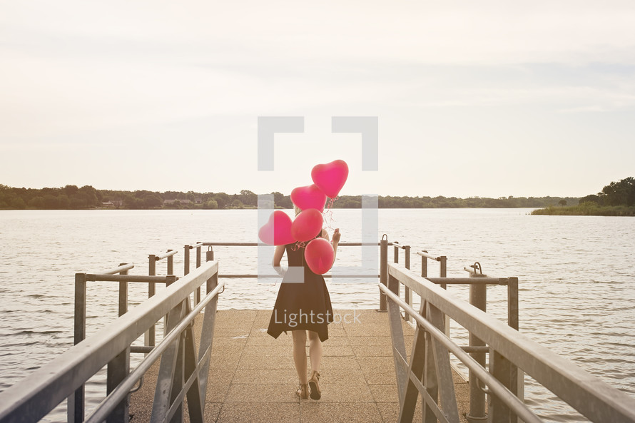 a woman walking on a dock carrying heart shaped helium balloons 