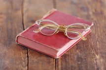 Eye glasses on an old red book which is on an rugged wooden table.
