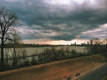 Street, river, and city skyline under storm clouds.