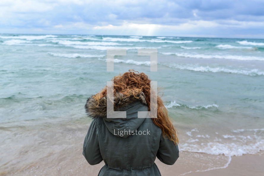 A woman in a coat stands at the ocean's edge.