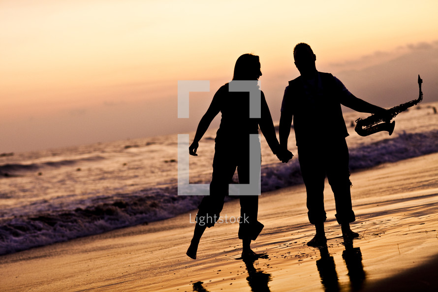 silhouette of a man and a woman holding hands on a beach holding a saxophone 