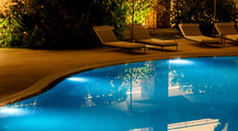 Luxurious swimming pool at night, beautifully lit, reflecting trees. Blue water sparkles under lights, creating a serene atmosphere for relaxation. Perfect for a tropical escape