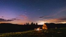 Friends around campfire by wooden hut in summer starry night sky with fast clouds Timelapse
