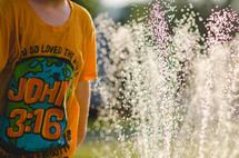 A child in a yellow John 3:16 t-shirt playing in a sprinkler on a sunny day.