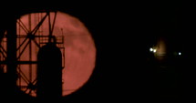 Red full moon rising from behind a large oil refinery.