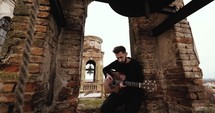 Man playing guitar in stone building with bell tower in the background