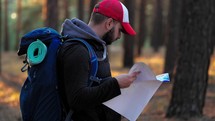 Hiking. Handsome bearded male tourist with backpack looking on map in forest. Lost hiker male with backpacks reading travel map, navigating and looking for directions in pine forest. Trekking concept.