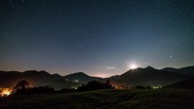 Starry night sky with moon rising over mountains in rural countryside Time lapse
