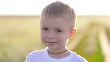 Closeup face of little boy looking to camera on the nature. Child boy smile in sunlight outdoors. Cute boy looking at camera while standing on sunset.