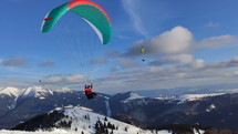 Men on green paraglider fly over snowy winter mountains.