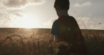 Young boy in a cape stands in a golden wheat field looking into the sunset