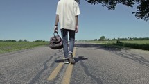 man walking down the middle of a rural road carrying a bag