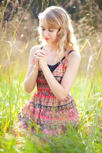 a blonde woman with praying hands kneeling in tall grass