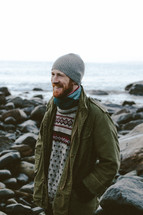 a man in a sweater, jacket, and beanie standing on a rocky shore 