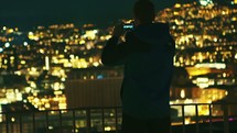 man on a balcony taking pictures of a city below at night 