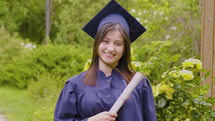 Smiling young woman holding diploma and wearing cap and gown outdoors looking at camera. Graduation concept. Slow motion 