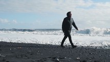 a man in a coat and boots walking on a beach in winter 