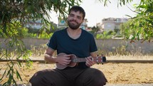 Smiling Man Plays Ukulele Musical Instrument In An Outdoor Park