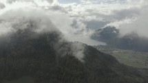 Aerial drone flying through the misty mountains with distant mountain lake and small buildings  below
