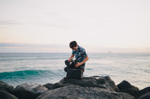 man near the ocean sitting on a rock looking into a bag 