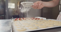 Baker sprinkling powdered sugar with a strainer onto cookies with strawberry jam.