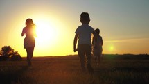Family concept. Mother and two children running through the meadow at sunset. Silhouette happy family during the travelling.