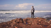 Man in hat walks on a rocky beach and looks at endless ocean, peaceful freedom travel background
