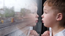 Close-up shot of a little curious boy looking out of the window in train. It's raining outside, child reflecting in the glass