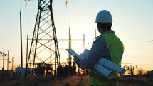 Power work concept. Engineer standing on field with electricity towers. Young engineer putting security helmet. Electrical engineer with high voltage electricity pylon at sunset background.