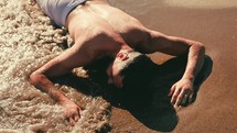 a man face down on a beach as water washes over him 