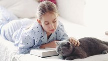Girl strokes a grey cat while reading a book. 