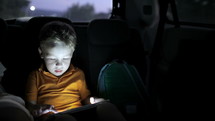 Little boy traveling on backseat of a car at night and using touch pad to entertain himself during the trip