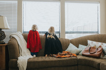 siblings in capes looking out a window standing on a couch 