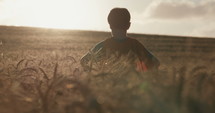 Young boy in a cape stands in a golden field during sunset - raising his hands in victory