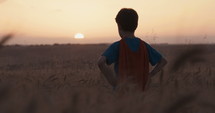 Young boy wearing a superhero cape stands in a golden wheat field looking into the sunset