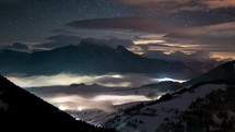 Magic starry night sky with stars in winter alpine mountains with foggy clouds in valley time lapse
