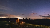 Sitting around the campfire with friends by wooden hut in peaceful summer evening forest nature under starry night sky Time lapse
