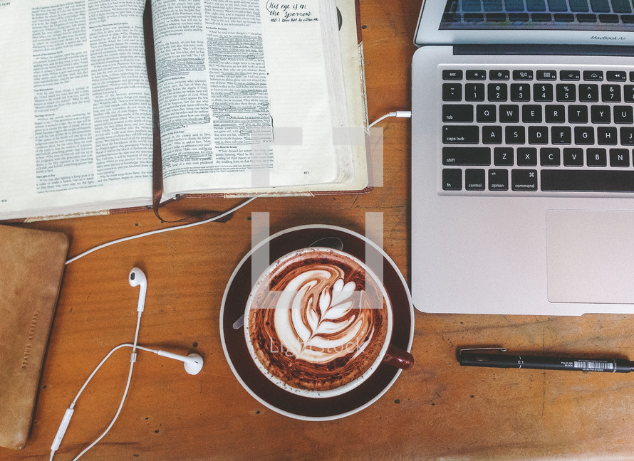 an open Bible, a laptop computer with earbuds, a pen, and a cappuccino 