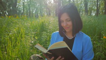 Young woman with brown straight hair reading book, smiling, enjoying sun and relaxing in park of forest, outdoors surrounded by trees. Sunny weather.
