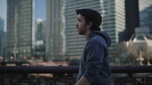 A contemplative young man walks along a bridge with city buildings in the background