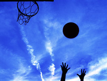 Silhouette of hands throwing basketball towards hoop with bliue sky in the background.